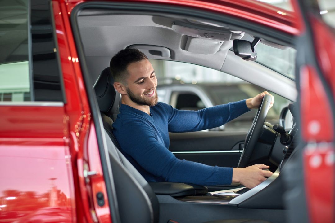 smiling man in a blue sweater sitting in the driver's seat of a red car with the door open