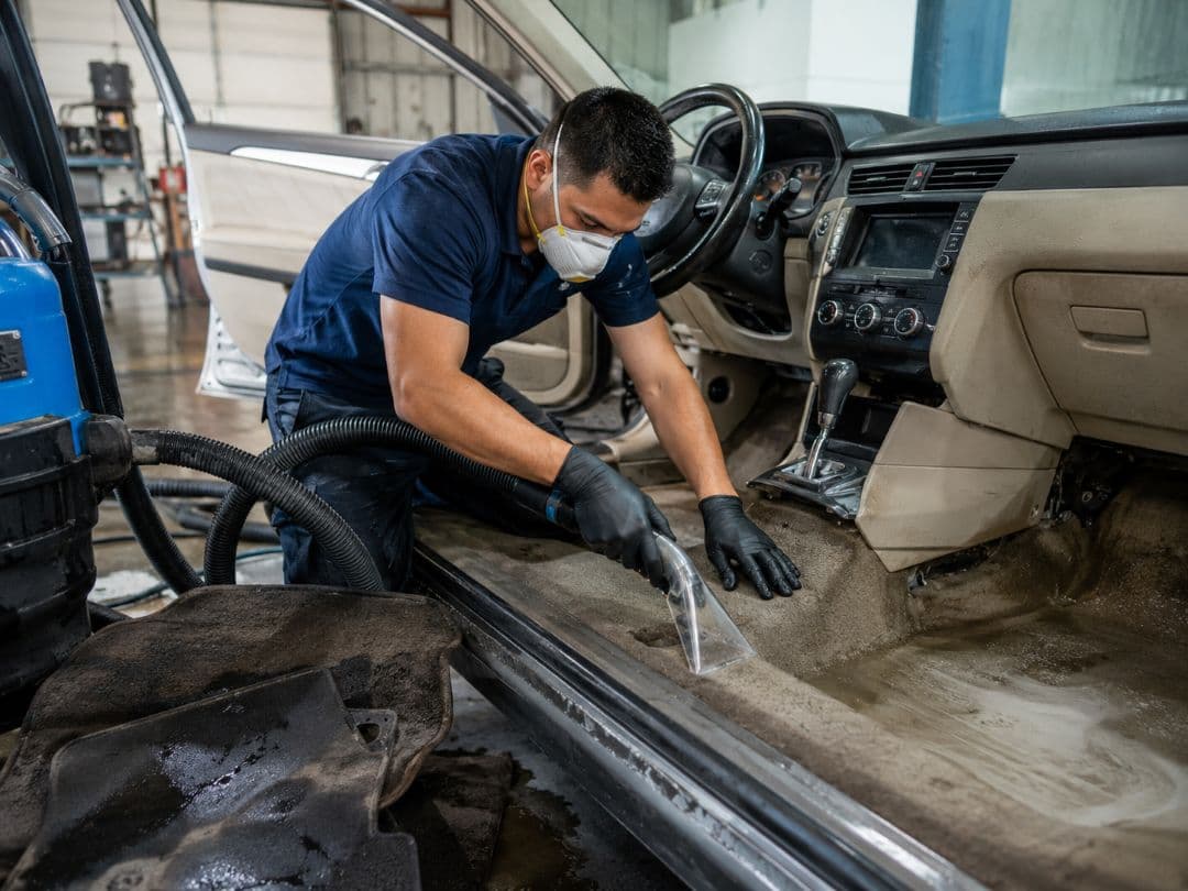 technician wearing a mask uses an extractor to clean muddy carpet inside a flood-damaged car