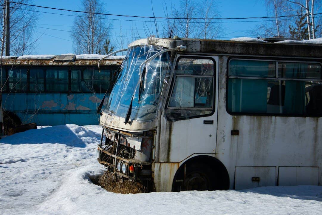 abandoned white bus with a damaged front end, broken windshield, and rusted body partially buried in snow