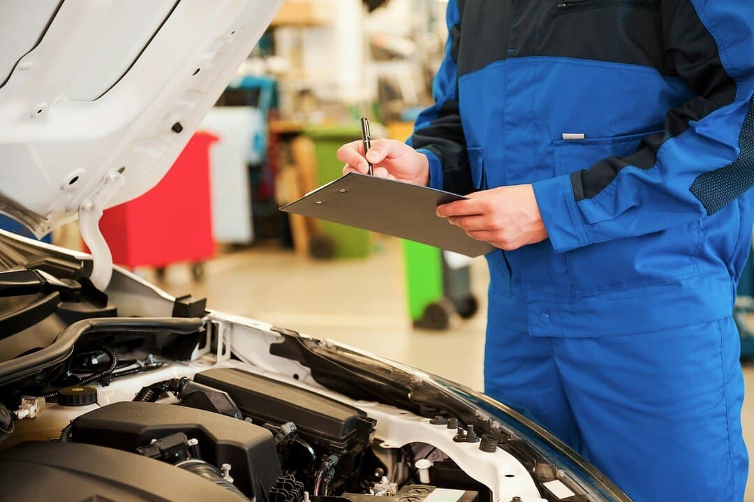 auto mechanic in blue workwear writing on a clipboard while inspecting a car engine with the hood open