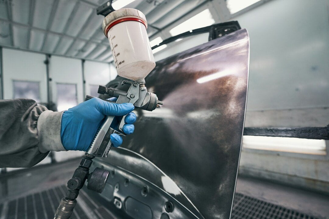 close-up of a worker wearing blue protective gloves using a spray paint gun to paint a car body panel