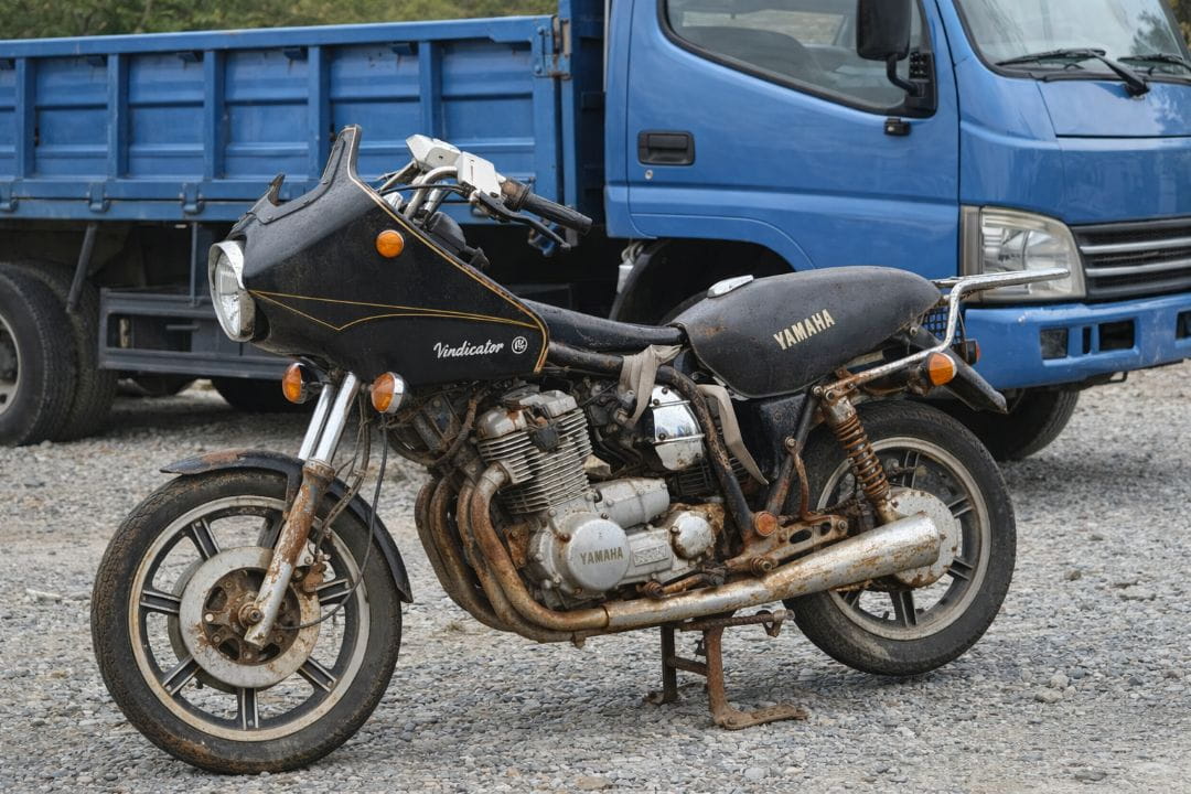 Una vieja motocicleta Yamaha oxidada estacionada sobre grava junto a una camioneta azul