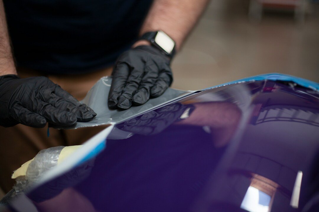 close-up of a worker wearing black protective gloves applying and smoothing a protective film onto a glossy purple car body panel